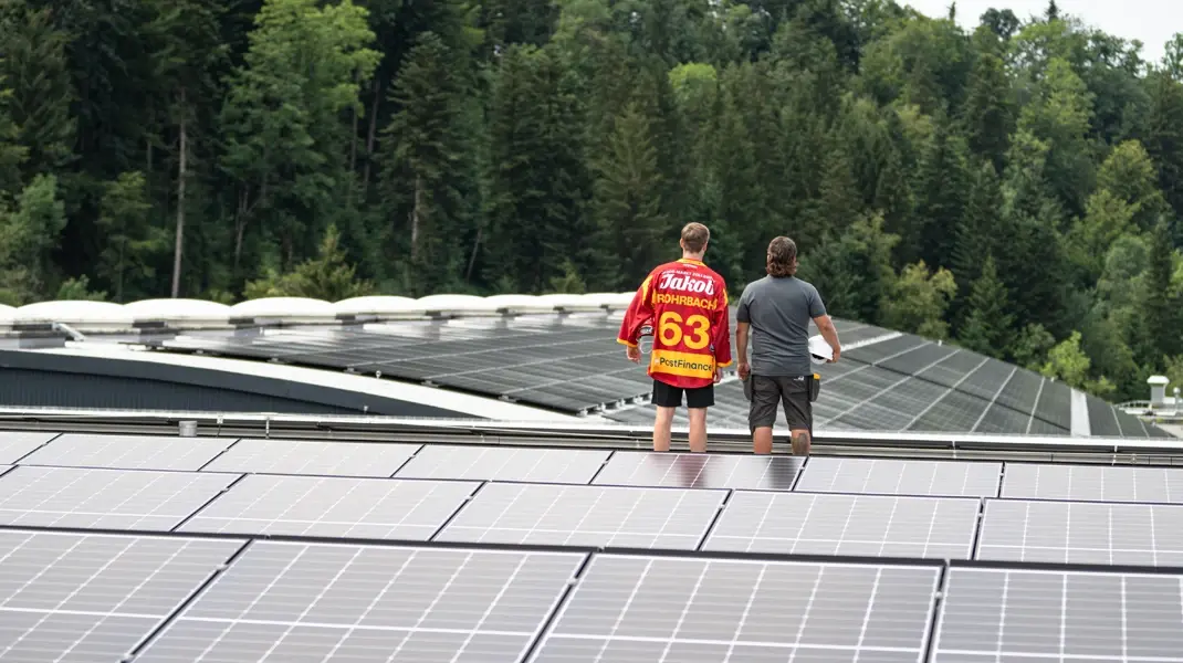 SCL Tigers Spieler steht mit Solarteur auf dem Dach und blickt auf die neue Photovoltaikanlage. Beide sind in ihren Arbeitskleider; einmal im Tigersdress und einmal in GLB Montur. 